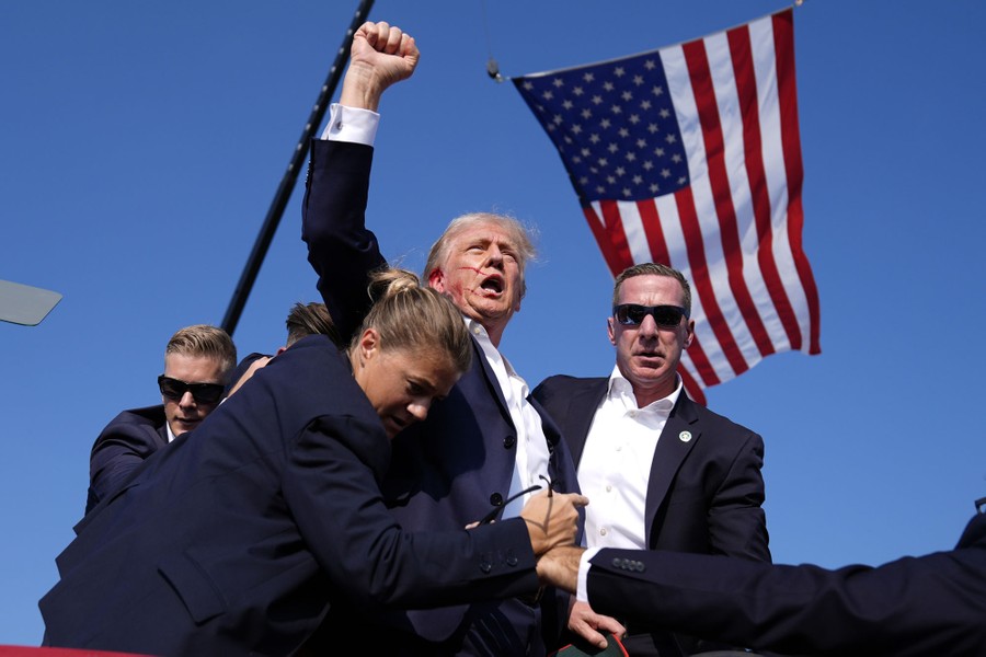 Former President Donald Trump is surrounded by Secret Service agents, backdropped by a large American flag, after an attempted assassination. Trump, with some visible blood on his face, raises his fist high.