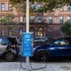 Two electric vehicles charging on a public charging station parked on a shady city street