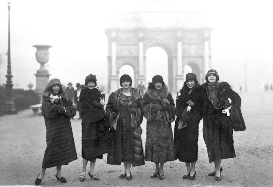 Six women wearing fashionable coats and hats pose in front of an arch in Paris.