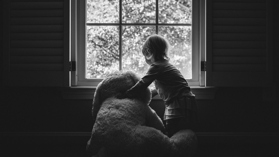 A young girl holding a large teddy bear stares out the window.