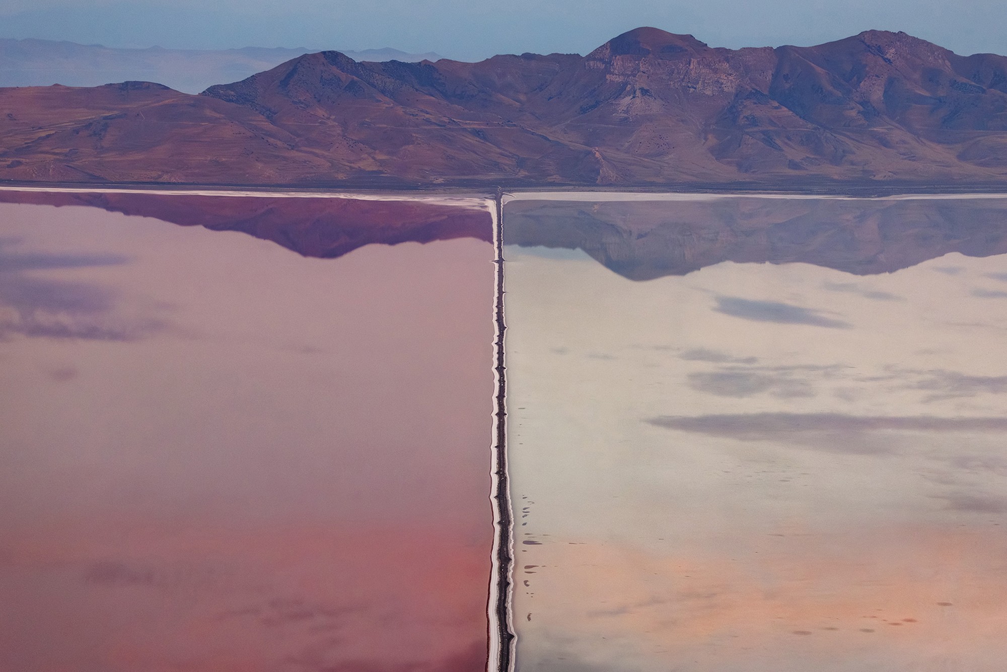 A view of a dividing dam in Utah’s Great Salt Lake, with two different bodies of water seen in varying hues of pink