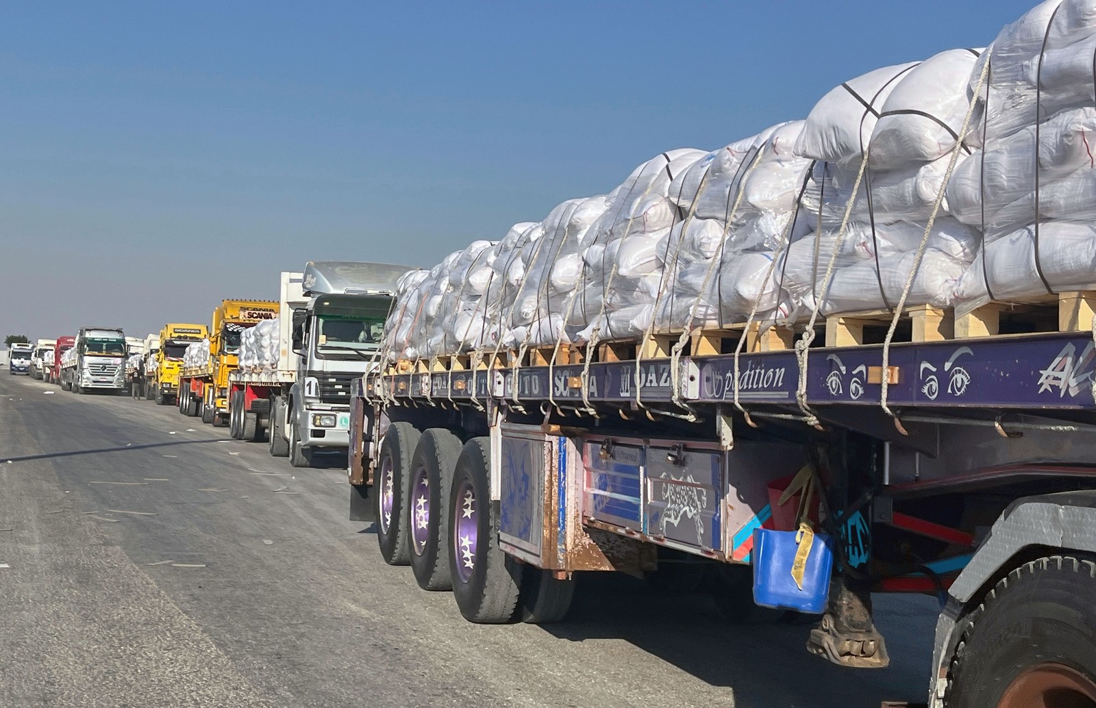 A long line of flatbed trucks on a road, each carrying bags of food aid