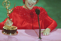 Woman in a red dress holding a golden Emmy trophy, smiling in front of a microphone.