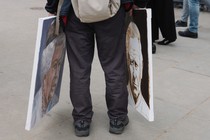 Someone shown from the legs down holds two paintings, one of Queen Elizabeth II and one of King Charles III