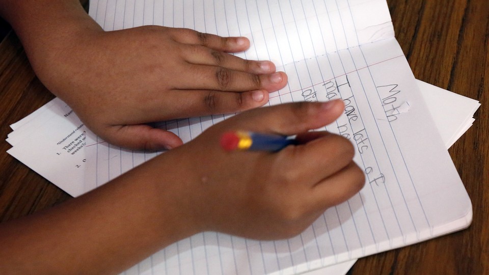 A child holds a pencil and writes on a sheet of notebook paper. 