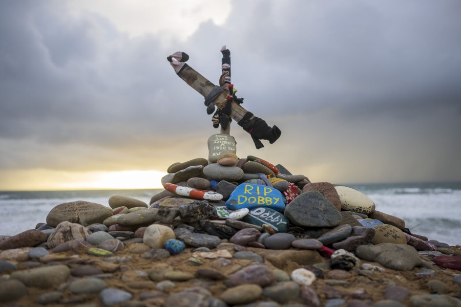 A crooked cross memorial stands on a beach, among a pile of painted rocks.