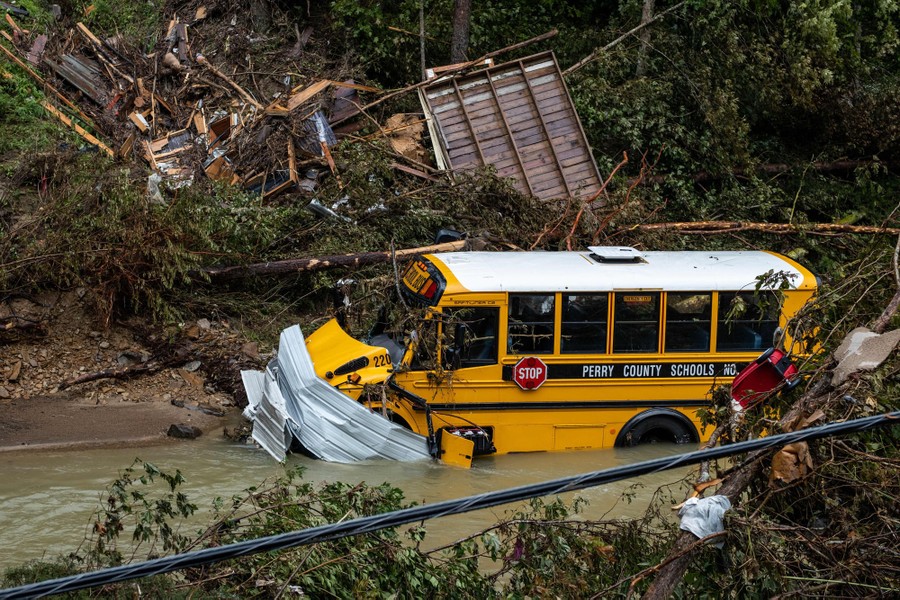A damaged school bus sits in a creek, surrounded by flood debris.
