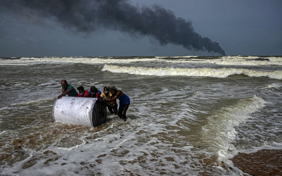 Five people push a large cylindrical object out of the surf, with black smoke rising from a point in the distance.