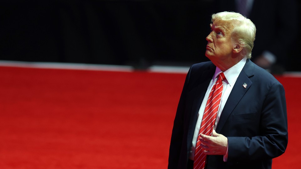 Donald Trump touches his tie and looks up while standing on a red carpet