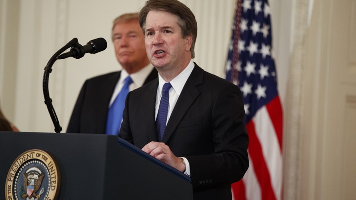 Brett Kavanaugh speaks at a podium with President Trump and an American flag in the background