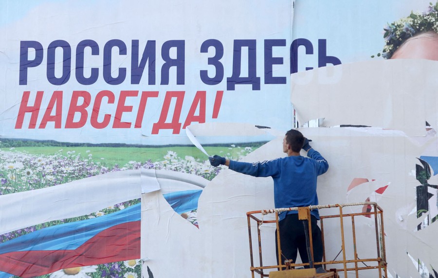 A worker tears away a billboard reading "Russia is here forever" in Russian.