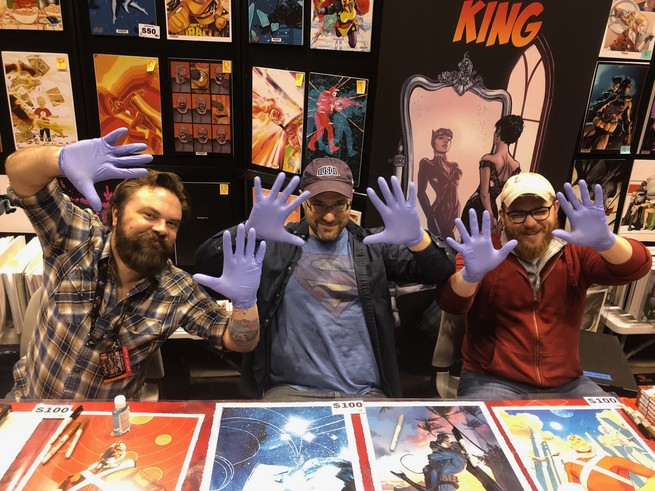 Three men sit at a table surrounded by prints of comic book artwork. They're wearing latex gloves and holding them up to the camera