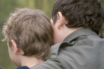 A father and son sit in a boat, their backs to the viewer. The father has his arm around the son.