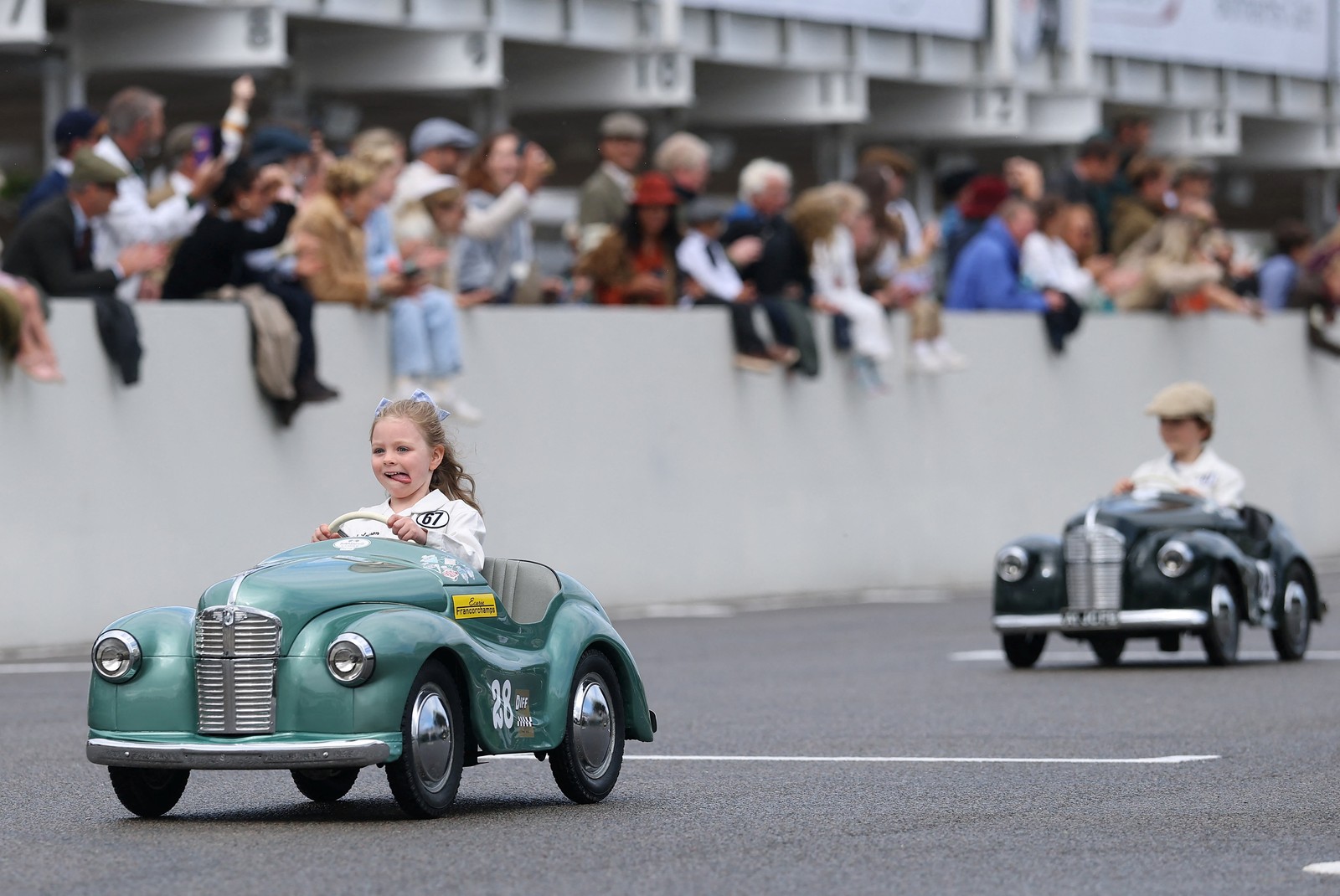 Children pass by a crowd, driving pedal cars in a race.
