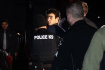 Photo of Luigi Mangione after his arrest looking back at the camera while being escorted away by a cop in a “Police K9” vest