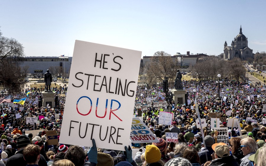 A crowd of demonstrators, chants and holds signs, including one that prominently says "He's stealing our future."