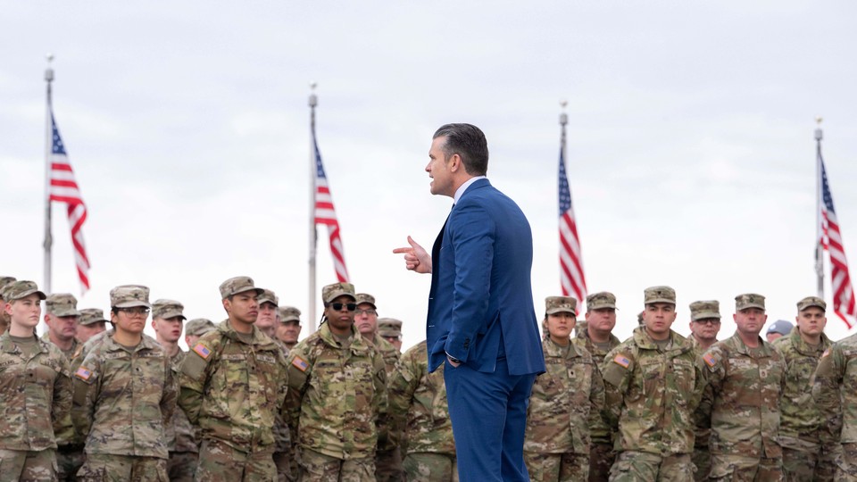 A color photograph of Pete Hegseth speaking in front of uniformed soldiers on a cloudy day; four American flags are seen in the background.