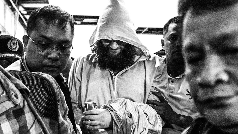 A hooded man with a beard holds a Coke can and keeps his head down. He is closely surrounded by half a dozen men who seem to be forcibly escorting him somewhere.