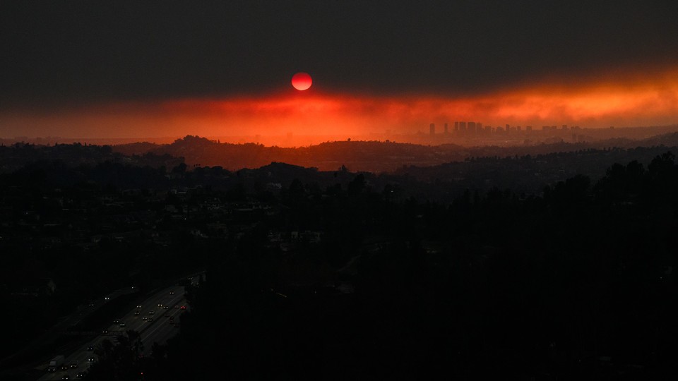 An aerial image showing vehicles driving on a freeway as the sun sets behind smoke from wildfires in Los Angeles
