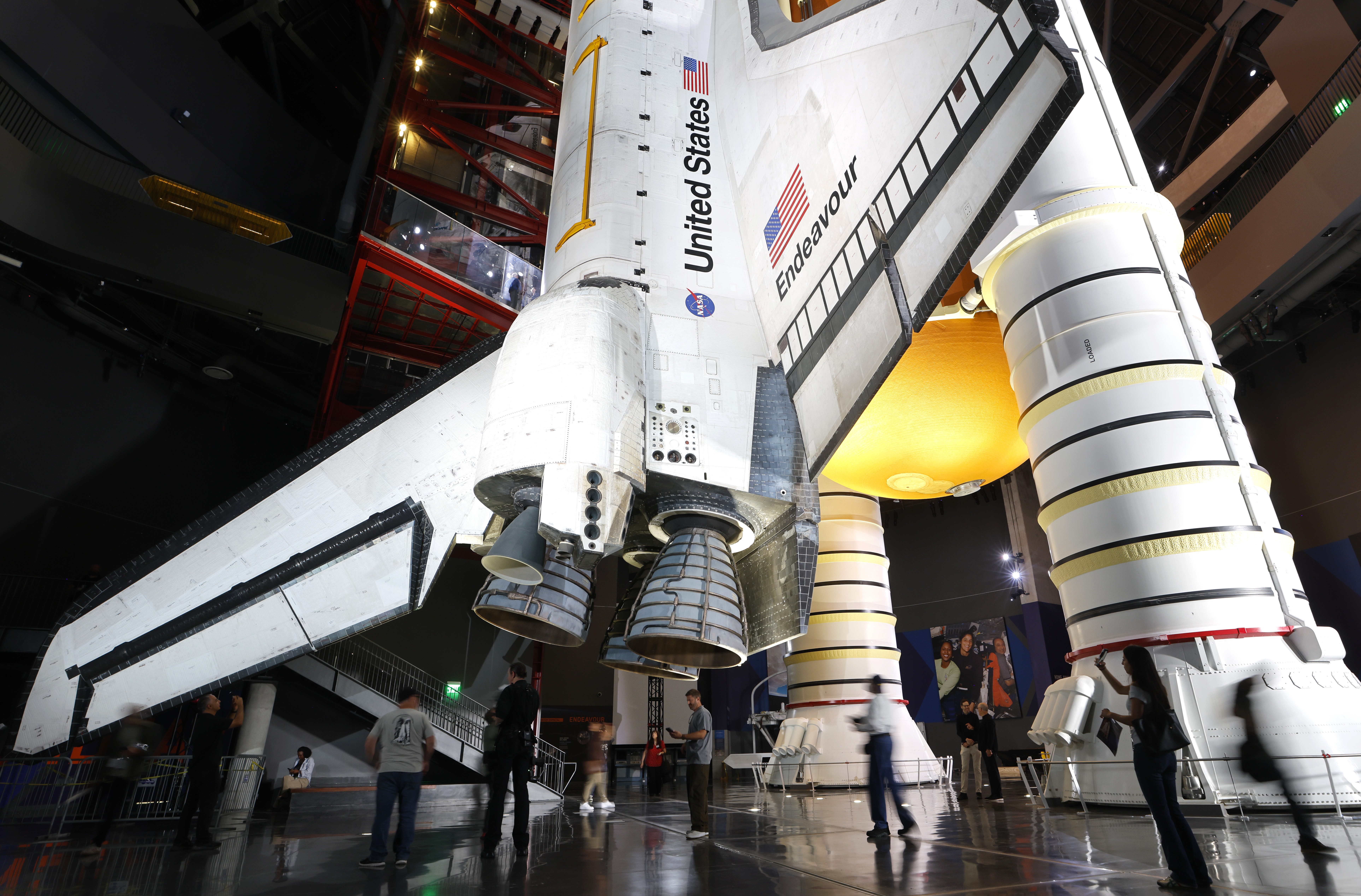 People walk near the base of a decommissioned space shuttle in a museum setting.