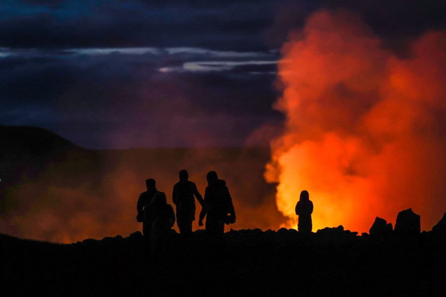 Several people stand and walk on rocks at night near a volcanic eruption.