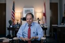 Framed by the U.S. and Alabama state flags, Doug Jones sits in an office.