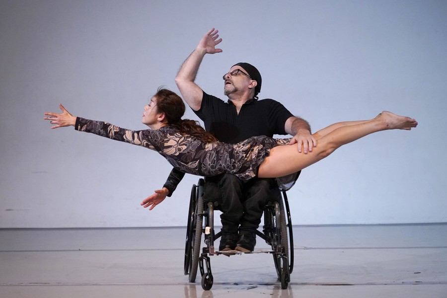 A pair of dancers, one in a wheelchair, rehearse on a stage.