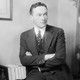 A black and white portrait of Walter Lippmann in a striped suit sitting on a writing desk in front of some books