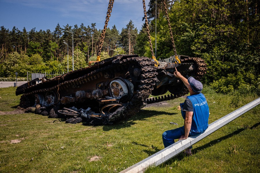 A person points up, sitting beside the remains of a tank, as it is lifted up by four large chains.
