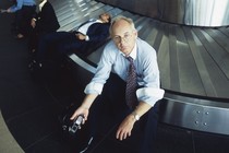 A man in a shirt and tie holds a cellphone and looks upset while sitting on a baggage carousel at an airport.