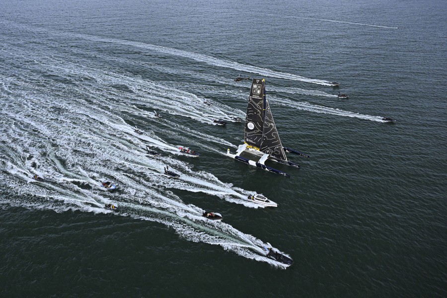 A racing trimaran sails, accompanied by many small boats.