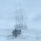 A small group of men walk across a snow-covered landscape toward the camera and away from an old ship.