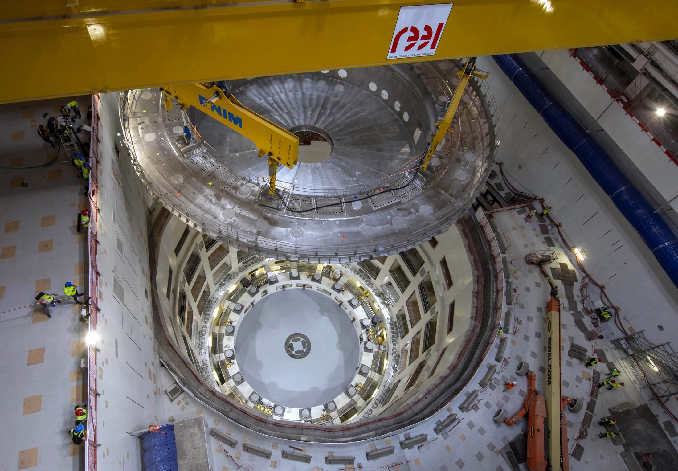 A large metal lid hangs from a gantry crane above a 30-meter-deep circular pit.