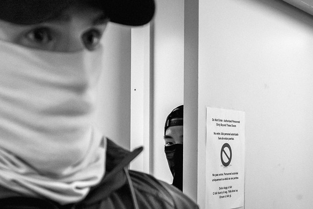 A black and white photographs of two ICE Officers, one in the foreground, and one peeking out from behind a door.