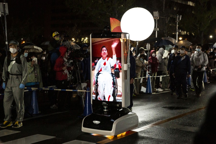 A robotic platform with a large video screen showing a torchbearer moves down a road, surrounded by onlookers.
