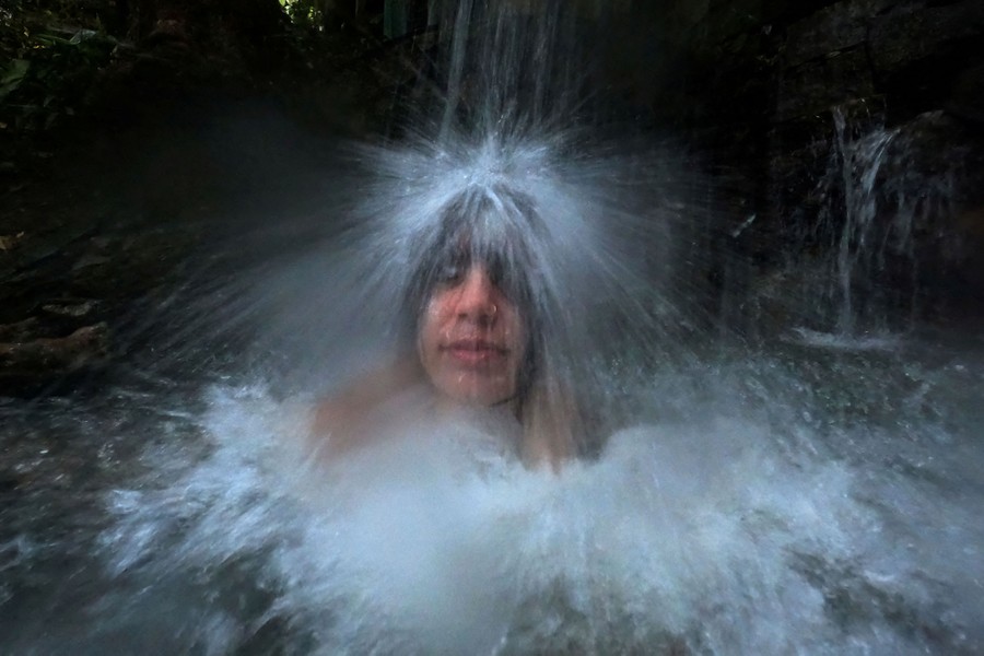 A person sits shoulder-deep in a pool in a stream, beneath a small waterfall that splashes on their head.