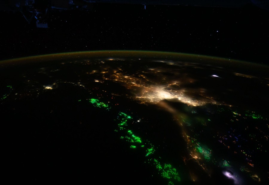 City lights and many green lights dot a coastline and just offshore, seen at night, from orbit.
