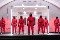Seven people wearing neon-pink jumpsuits and black masks with different symbols