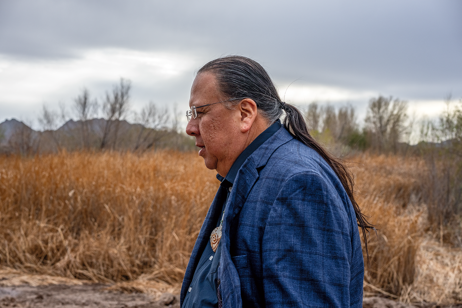 photo of side view of man in glasses with long dark ponytail wearing blue blazer with dry grasses and mountain ridge in background