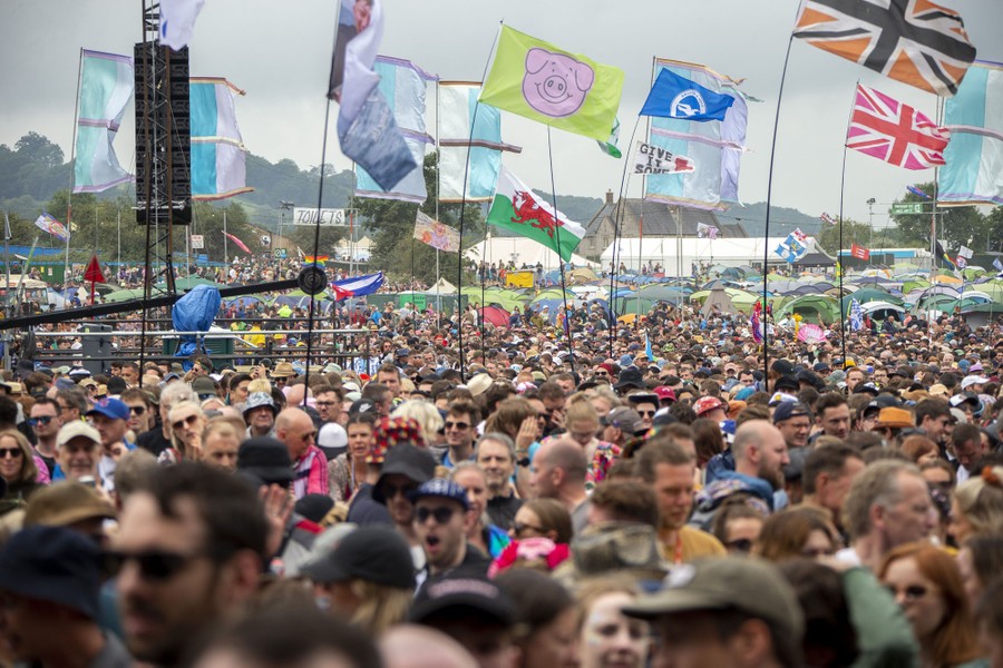 A large crowd moves across a field, with banners and tents seen in the background.