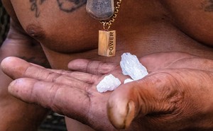 Close-up view of 3 bright-white crystal rocks resting in a man's palm in front of his bare tattooed chest and necklaces, one with a rectangular pendant with the word "POWER"