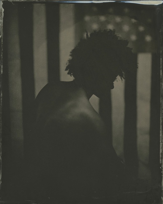 A black man is silhouetted by an American flag in a black and white tintype photo