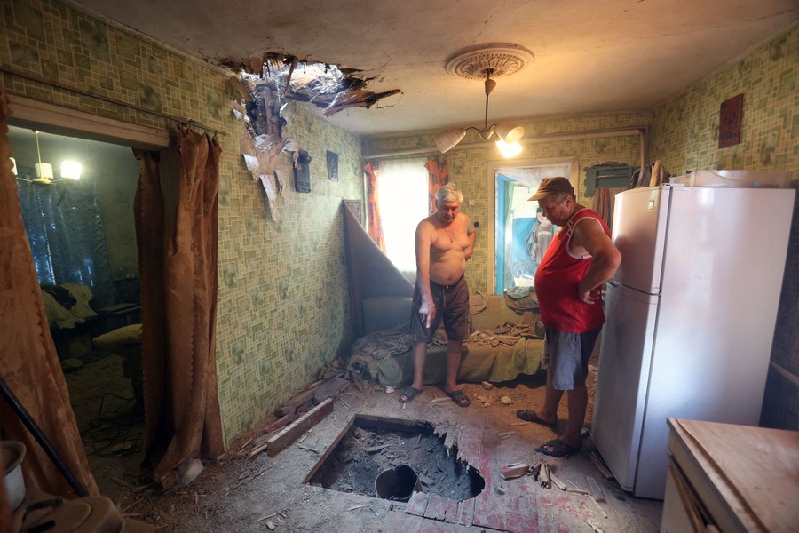 Two men stand in a kitchen looking at holes punched through the floor and ceiling.