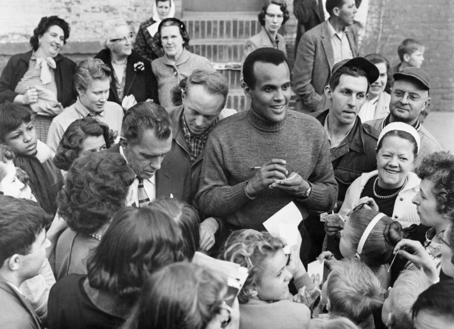 Harry Belafonte and Ed Sullivan stand surrounded by fans, signing autographs.