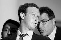 A black and white photograph of a bespectacled man whispering into Mark Zuckerberg's ear