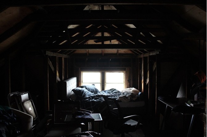A dimly lit picture of an attic with a beamed ceiling, and an unmade bed tucked into a small nook next to three tiny windows