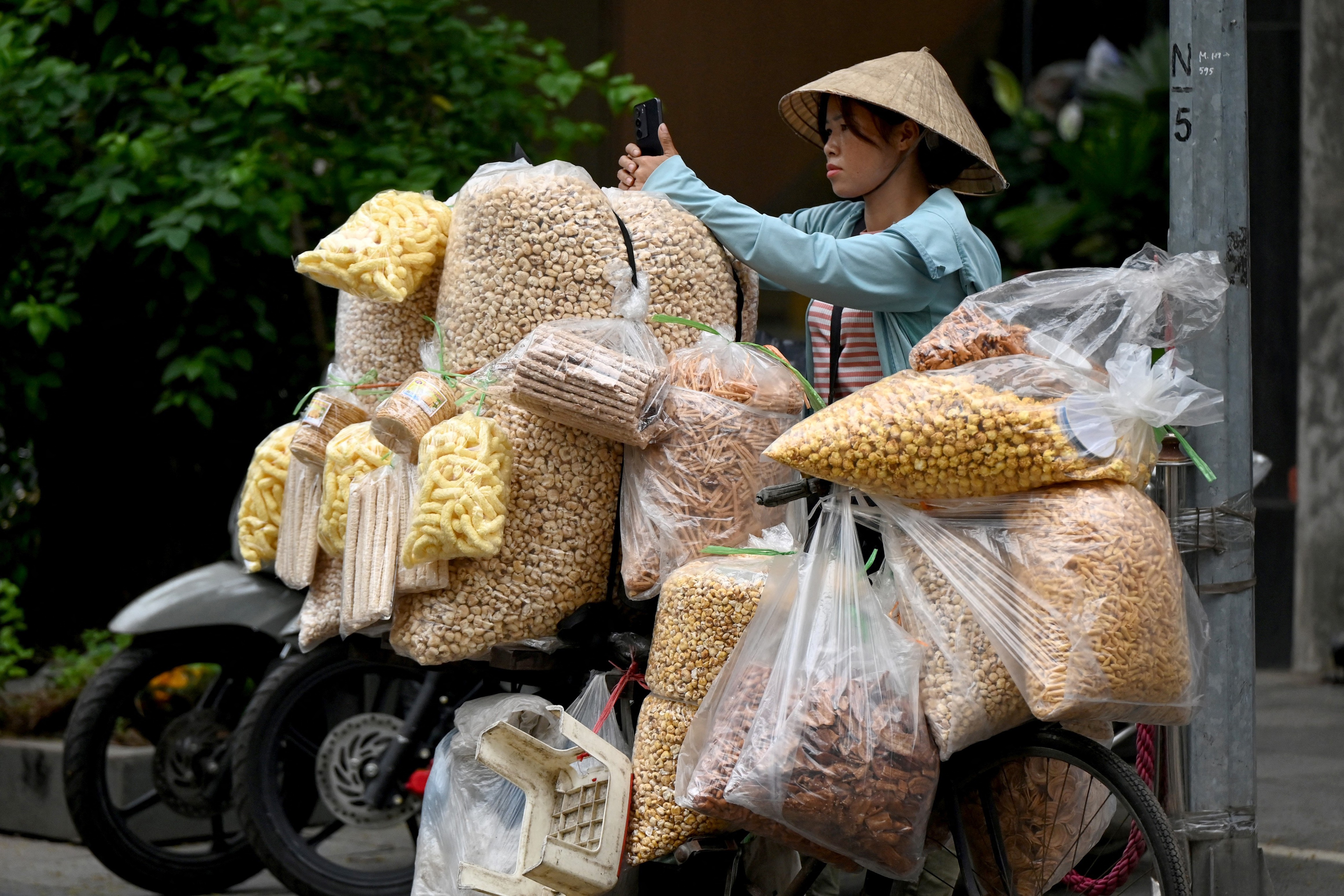 A street food vendor uses a mobile phone while waiting for customers on a scooter that is heavily loaded with clear plastic bags full of snacks.