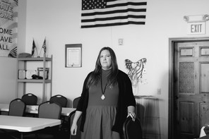 A black-and-white photo of a white woman standing in a room with the American flag behind her