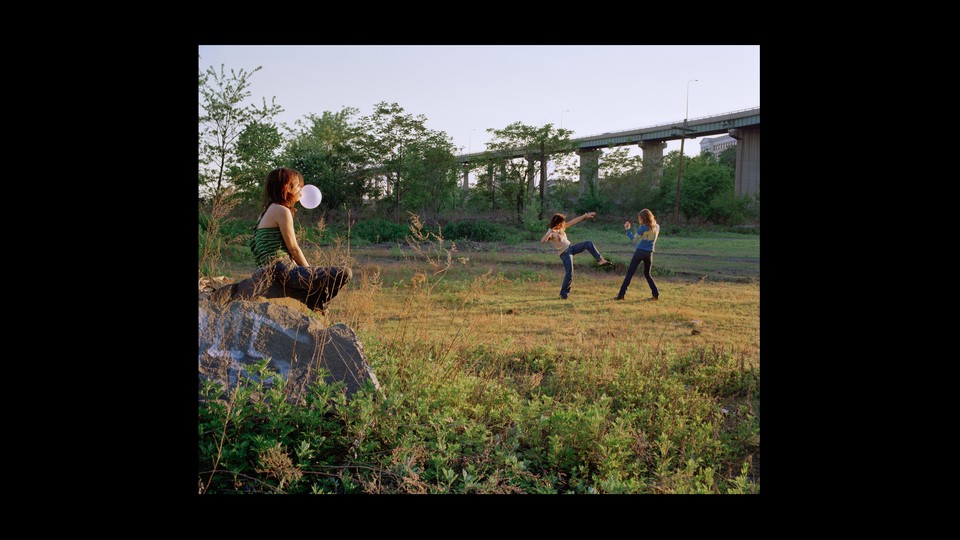 A girl sits on a rock blowing a gum bubble, while two girls in the distance play fight on a green field under a highway.