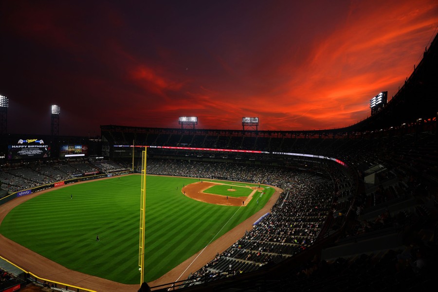 A sunset illuminates clouds above a baseball stadium during a game.